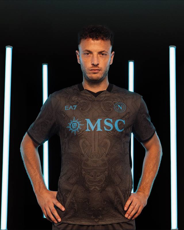 Amir Rrahmani posing in the SSC Napoli 2024/25 third shirt, which features a dark grey design with baroque-inspired graphics and light blue details.