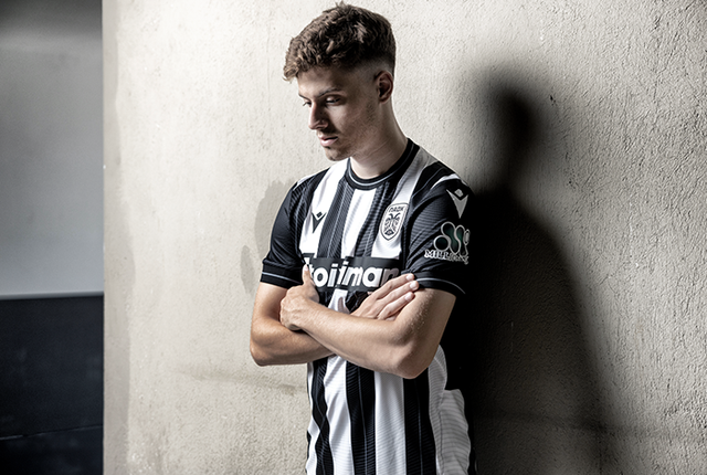 A model poses in the PAOK FC 2024/25 home shirt against a concrete wall.