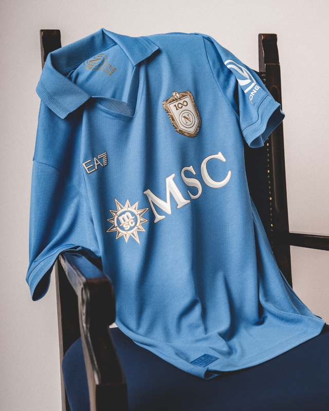 The SSC Napoli home shirt is presented draped over a dark wooden chair.