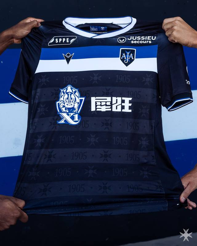 A close-up of the AJ Auxerre away shirt by Macron, featuring a navy blue design with a horizontal blue and white stripe across the chest.