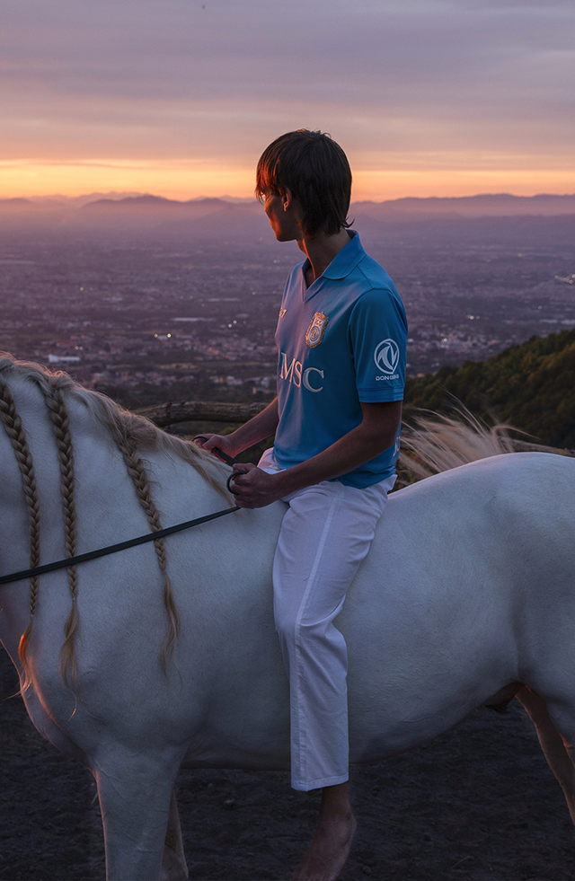 A campaign photo showing a model wearing the SSC Napoli home shirt while riding a white horse at sunset.