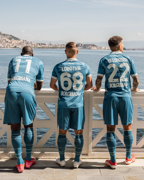 Three SSC Napoli players wearing the Partenope fourth kit are shown from behind, leaning against a white railing overlooking the sea.