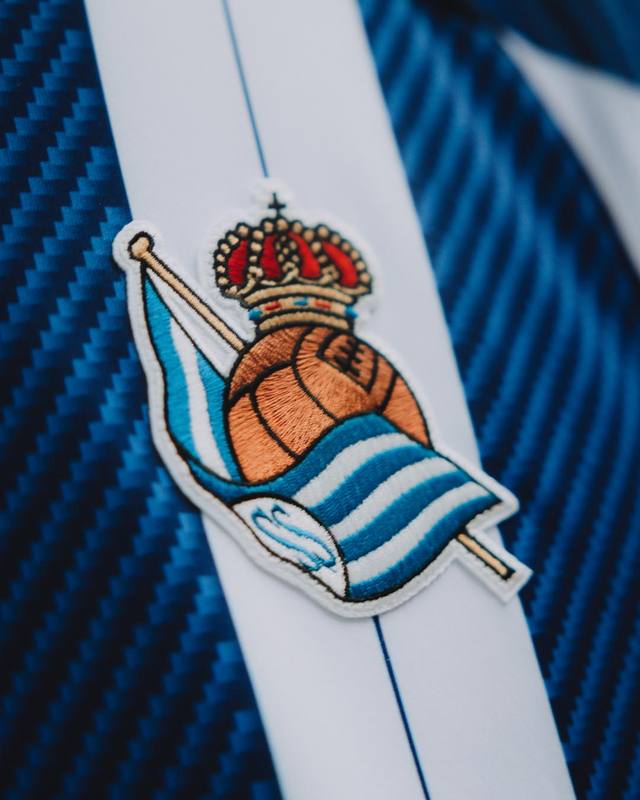 A close-up detail of the Real Sociedad club crest on the blue and white striped home shirt.