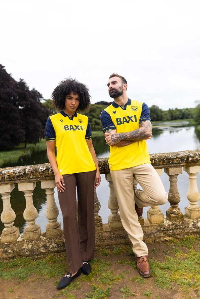 A promotional photo of two models posing in the Oxford United FC 2025/26 home shirt by a scenic river bridge.