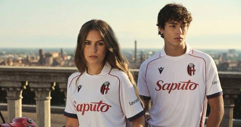 Two models wearing the white Bologna FC 1909 away kit pose in front of a city skyline.