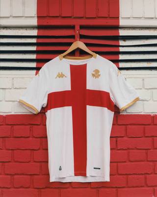 A white Genoa away shirt featuring a red St. George's cross, hanging on a wooden hanger against a red and white brick wall.